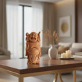 A hand-carved light brown wooden statue of the Laughing Buddha with a wide smile, holding a staff over his shoulder with a small child and a fish.